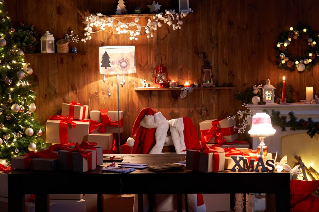 Santa Claus costume and hat hanging on chair at table with Merry Christmas decor gifts presents on holiday eve in cozy Santa home workshop interior late in night with light on xmas tree and fireplace.